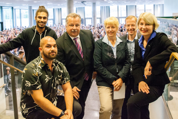 Die 6 Personen stehen nebeneinander und schauen in die Kamera. Im Hintergrund sind die neuen Studierenden zu sehen.