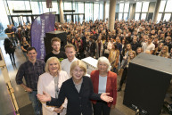 Drei Frauen und drei Männer stehen vor einer großen Menschenmenge und lächeln in die Kamera