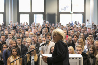 Eine Frau am Mikrophon steht vor einer großen Gruppe von jungen Leuten