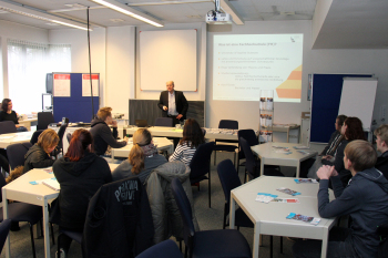 Mann steht neben Leinwand mit Präsentation, vor ihm sitzen Leute an Tischen.