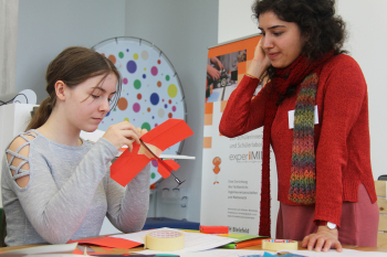 Ein Mädchen hält einen roten Papierflieger in der Hand und neben ihr steht eine Frau