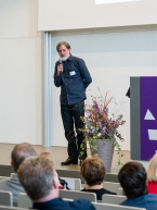 Ein Mann auf einer Bühne mit Mikrofon in der Hand neben ihm steht ein großes Blumengesteck