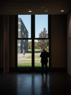 Ein Mann steht vor einem großen bodentiefenfenster blickt hinaus auf ein Gebäudeteil einen Baum und einen jungen Mann der mit dem Rücken zu ihm vor dem Gebäude steht