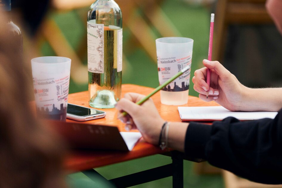 Auf einem Bierzeltgarnitur-Tisch stehen Plastikbecher und eine Flasche Wein. Hände halten Bleistifte in der Hand.
