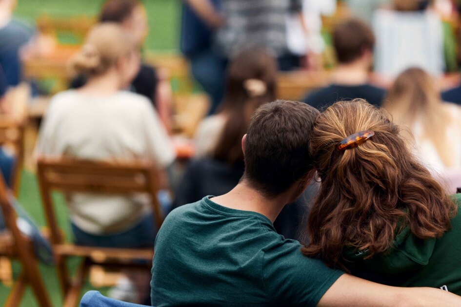 Zwei Personen sitzen nebeneinander, die eine hat den Arm um die andere Person gelegt..