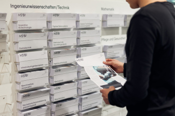 Ein junger Mann steht vor der Flyerwand mit den Studiengangsflyern der HSBI und schaut sich einen Flyer an