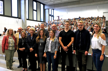 Eine Personengruppe steht in einem Hörsaal, dahinter sitzen Studierende.