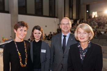 Drei Frauen und ein Mann vor der Bühne im Audimax mit dem Publikum im Hintergrund
