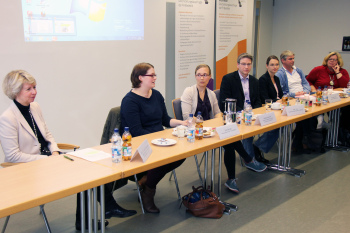 Sieben geschlechtsgemischte Personen sitzen nebeneinander an Tischen und referieren in der FH Bielefeld zu Studieren mit Kind.
