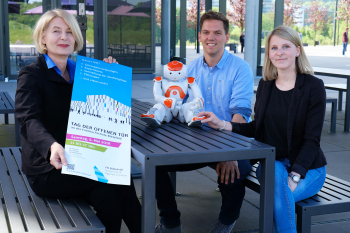 Zwei Frauen und ein Mann in der Mitte sitzen draußen an einem Tisch mit dem Plakat zum Tag der offenen Tür in der Hand.