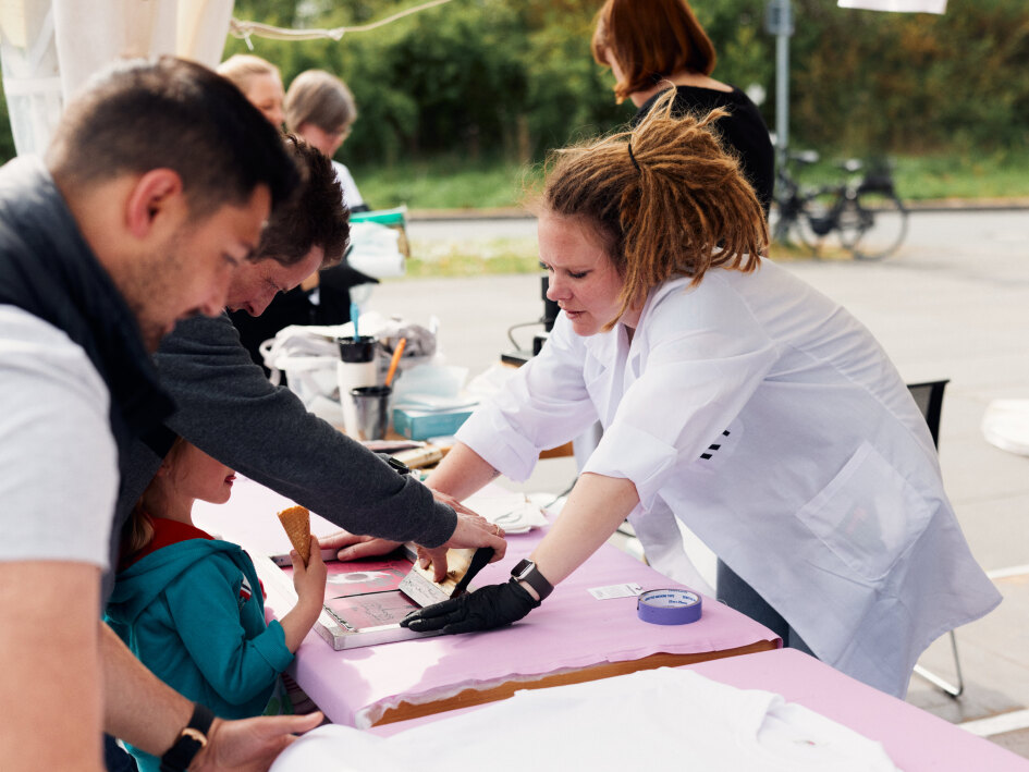 Eine Frau in einem Kittel assisitert Besuchern beim Siebdruck