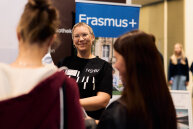 Eine Frau in einem HSBI Tshirt spricht mit zwei Besucherinnen im Hintergrund sieht man ein Erasmus+ rollup