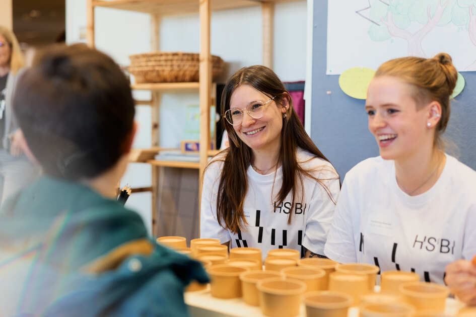 Foto: Zwei Studentinnen sitzen an einem Tisch mit vielen kleinen Bechern und sprechen mit einem Kind