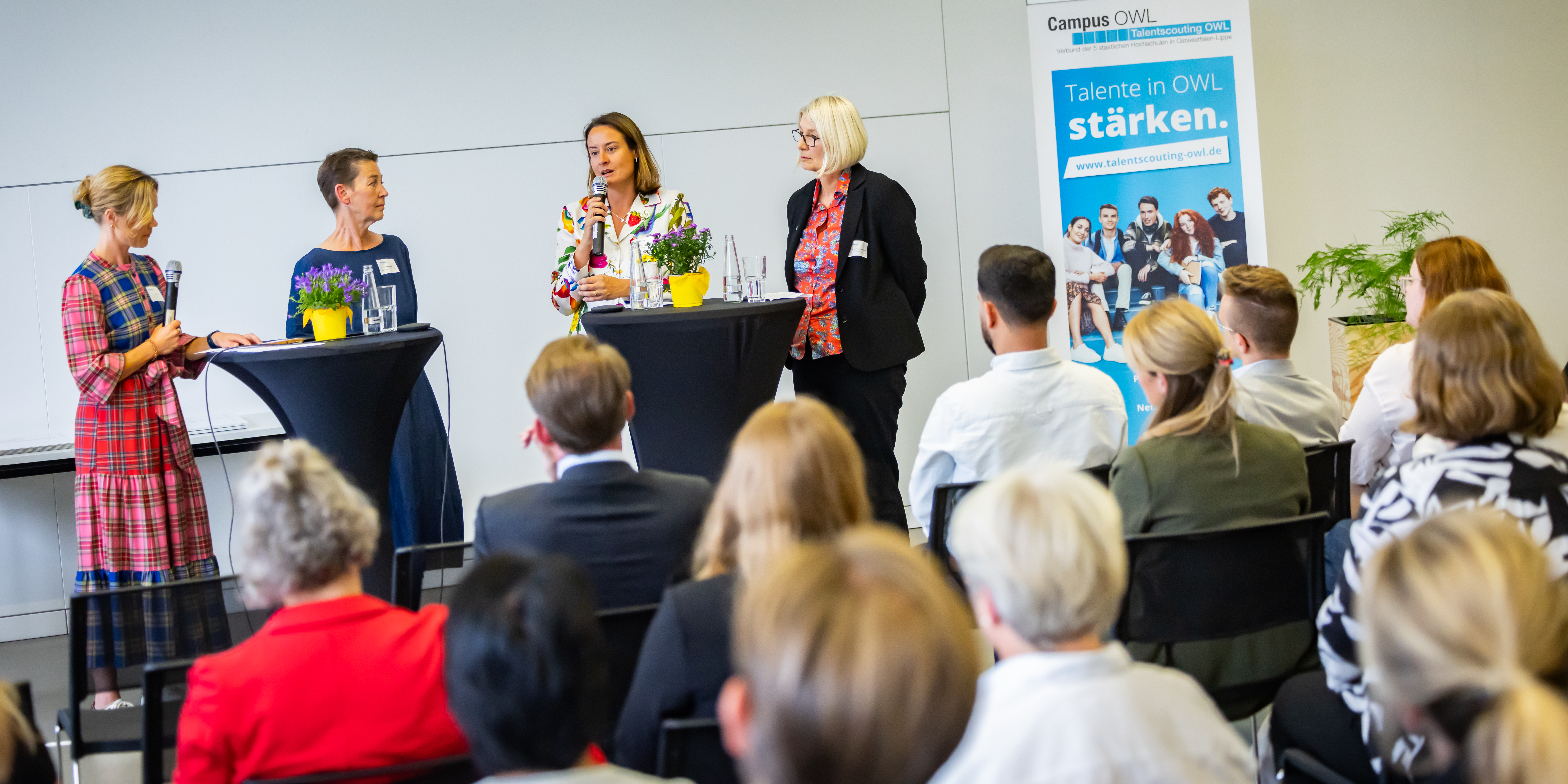 Vier Frauen stehen an Stehtischen im Konferenzbereich der HSBI vor Publikum