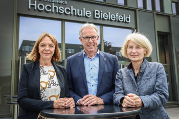 Drei Personen stehen an einem Stehtisch unter dem schriftzug Hochschule Bielefeld am Hauptgebäude der HSBI