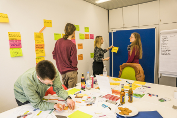 Vier Personen stehen in einem Raum an einer Pinnwand. An den Wänden hängen bunte beschriebene Zettel