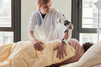 Eine Pflegekraft mit einer Arm-Orthese lagert eine im Krankenbett liegende Person um