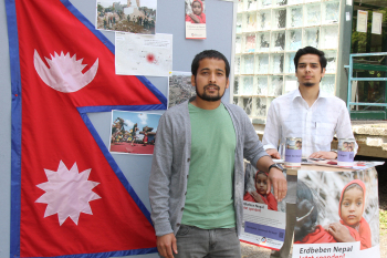 Die beiden Studenten stehen neben einem Stehtisch und eben einer Stellwand mit der nepalesichen Flagge sowie Bildern aus dem Erdbebengebiet.