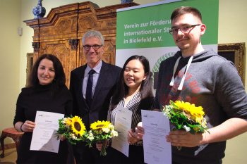 Vier Personen stehen vor einem Banner und einem alten Schrank. Sie halten Blumensträuße und Urkunden in der Hand.