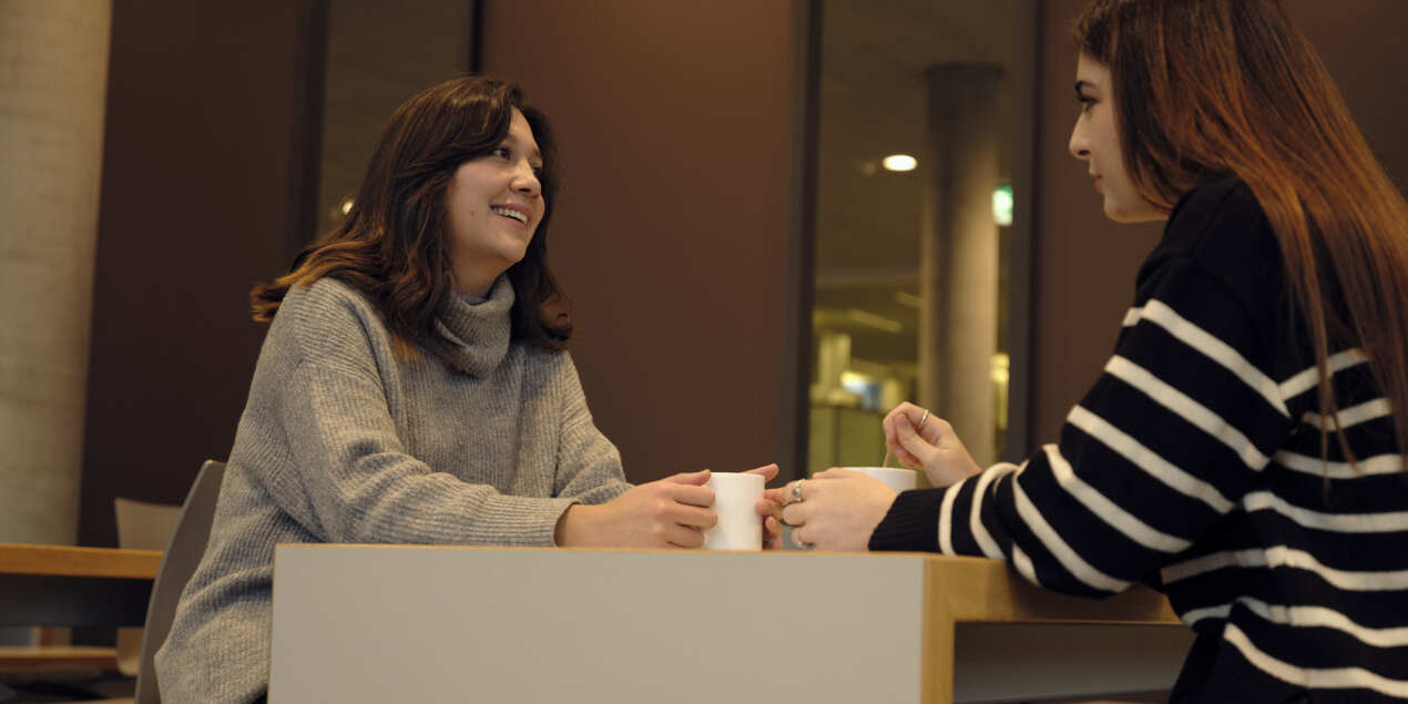 Ana Rangel und Chiara Gallizia sitzen an einem Tisch in der Cafeteria der HSBI und unterhalten sich Vor beiden steht eine weiße Tasse mit Kaffee Rangel schaut Gallizia an und lächelt dabei