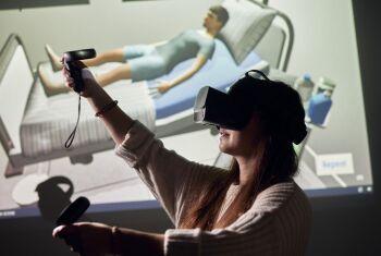 A woman wearing VR glasses is standing in front of a projection screen showing a simulation of a hospital bed