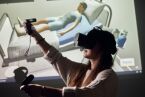 A woman wearing VR glasses is standing in front of a projection screen showing a simulation of a hospital bed