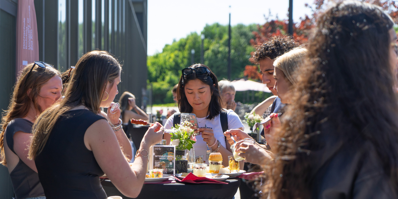 Fünf Personen stehen um einen Tisch unterhalten sich und essen gemeinsam