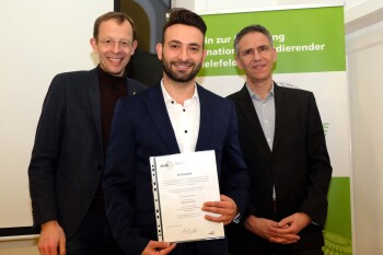 Drei Männer stehen nebeneinander, der Mann in der Mitte hält eine Urkunde in den Händen