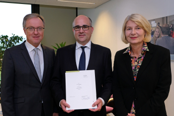 Drei Personen stehen nebeneinander und blicken in die Kamera, die mittlere Person hält eine Urkunde.