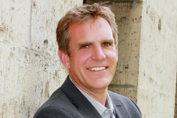 Ein Mann im Anzug steht vor einer Steinwand und lächelt in die Kamera