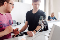 Zwei Personen betrachten eine Versuchsanordnung, die am Arm eines Studierenden angebracht ist