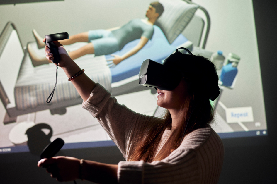Eine Studentin mit einer VR-Brille vor einem Monitor, auf dem ein Pflegezimmer zu sehen ist