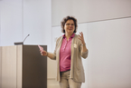 Beim Makeathon des InCams@BI Projekts steht Heike Wulf neben einem Rednerpult und hält einen Vortrag Sie hat mittellange braune Locken und trägt eine graue Strickjacke darunter hat sie ein rosa Poloshirt