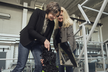 Marion Hövelmeyer und Eva Schwenzfeier-Hellkamp stehen in einer Halle hinter einem Trichter sie geben dunklen Kunststoff hinein.