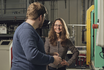 Links ist ein Mitarbeiter aus dem Bereich Kunststoffforschung von der Seite zu sehen er unterhält sich mit Christiane Nitschke die neben einem Gerät in der Experimentierhalle steht.