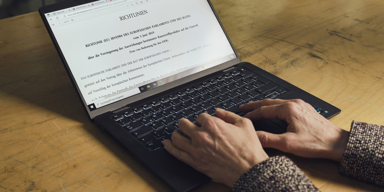 Auf einer Holzoberfläche liegt ein aufgeklapptes Laptop Auf der Tastatur tippen zwei Hände etwas ein Auf dem Bildschirm ist ein Richtlinientext zu sehen