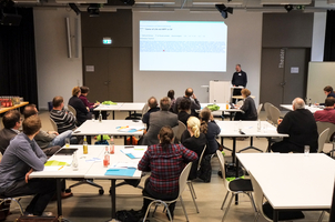 Seminarsituation, Referent steht neben der Leinwand, Menschen sitzen auf Stühlen an Tischen.