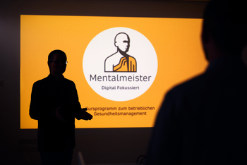 Auf orangen Hintergrund wird das Logo von Mentalmeister auf eine Wand projeziert