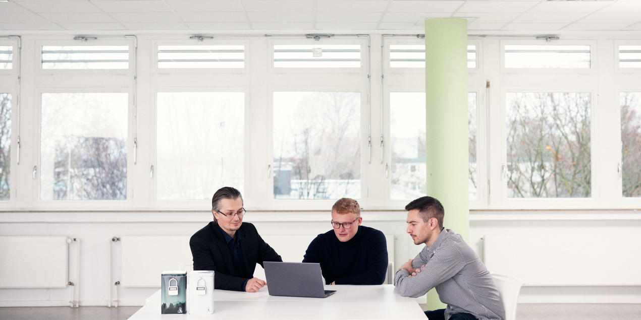 Lukas Gawor, Maik Abrams und Tobias Patzer sitzen in einem hellen Raum an einem Tisch und schauen auf ein Laptop