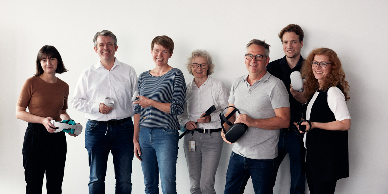 Gruppenbild der am Virdipa Projekt beteiligten Personen die vor einem hellen Hintergrund stehen und unterschiedliche VR Brillen und Controller in der Hand halten
