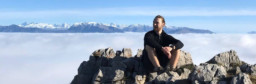 Moritz Mey sitzt auf einem Felsen, im Hintergrund eine Wolkendecke und eine Bergspitze