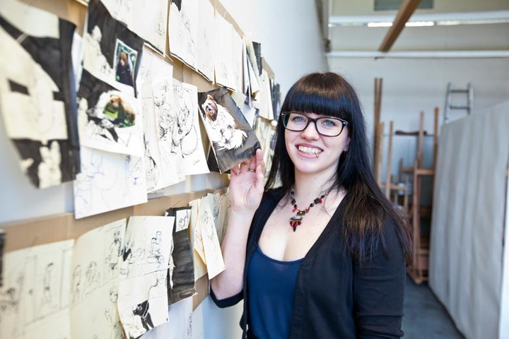 An eine Wand sind viele unterschiedliche Zeichnungen und Fotokollagen geheftet. Eine Studentin hält eins der an der Wand befestigten Blätter in der Hand, zeigt es und lächelt dabei in die Kamera.